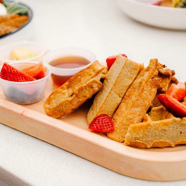 Waffles and French toast sticks with strawberries, syrup, and butter on a wooden tray.