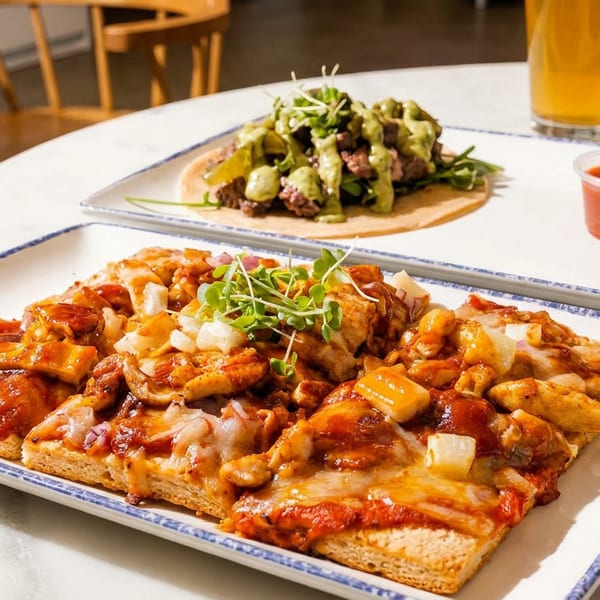 Flatbread pizza with chicken and pineapple, a taco, and a glass of beer on a white table.