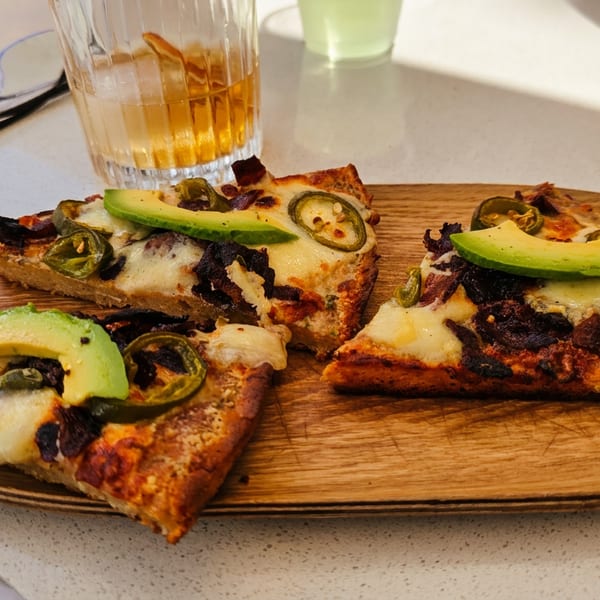 Three slices of pizza with avocado and jalapeños on a wooden plate, with a drink in the background.