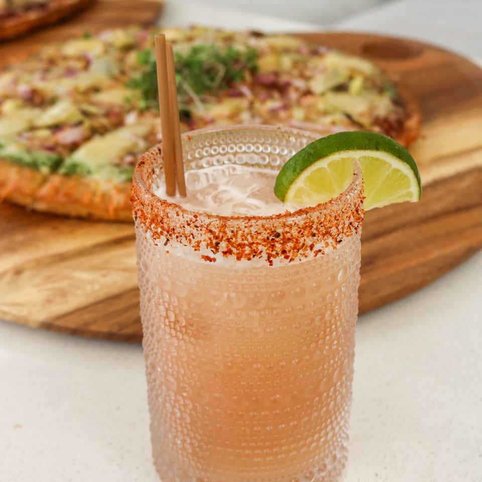 Lime-garnished drink with a seasoned rim beside pizza.